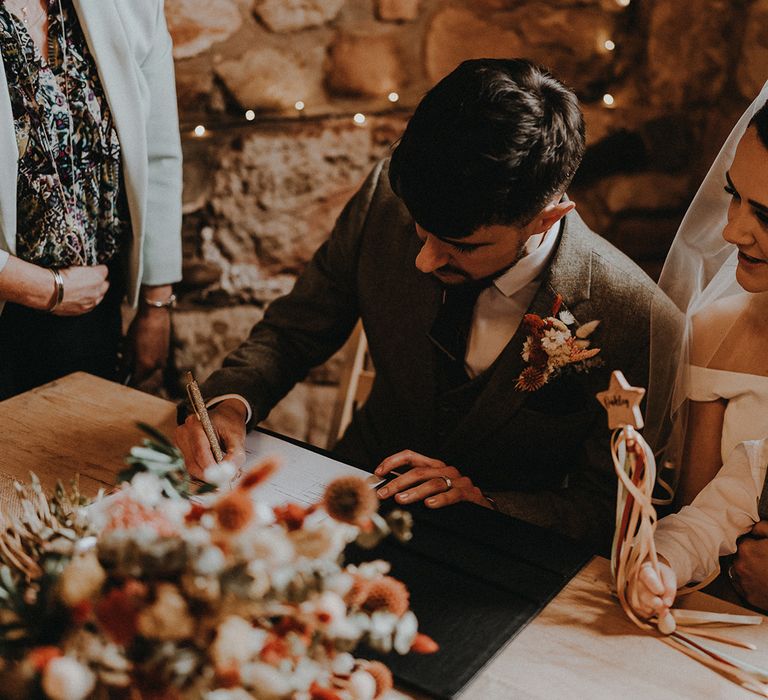 Bride and groom sit together to sign the wedding register at their rustic barn wedding 