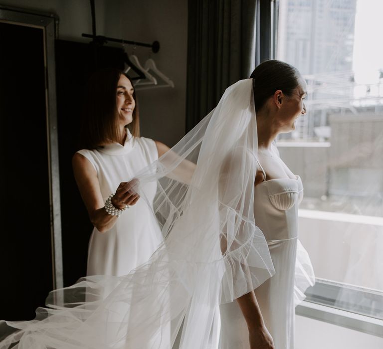 Mother of the bride helps the bride get ready in her satin wedding dress and ruffle detail wedding veil 