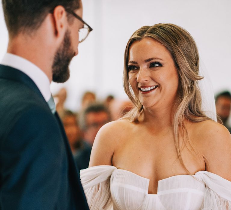 Bride in corset style wedding dress by Alena Leena with half up half down wedding hair smiling at the groom during their wedding ceremony 