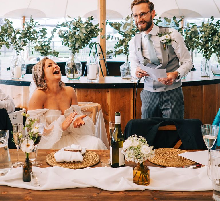 Groom in white shirt and grey waistcoat stands up to deliver his wedding speech with the bride clapping and laughing along