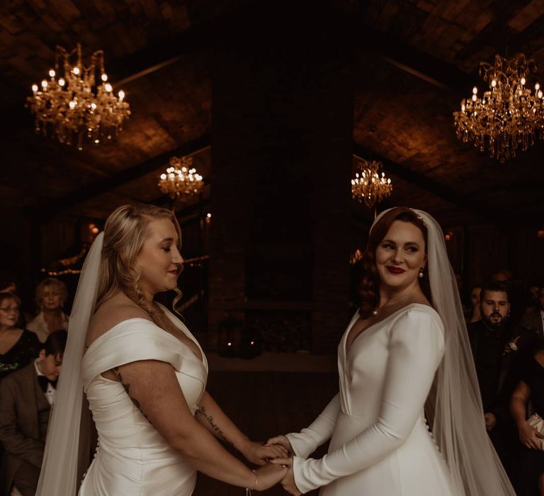 Bride in satin short sleeve deep v wedding dress and church length veil holding hands with bride in long sleeve deep v wedding dress and church length veil at the alter of Hidden River Cabins