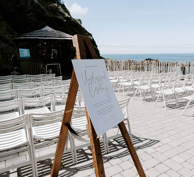 White and gold minimalist wedding welcome sign on wooden easel at Tunnels Beach looking over the sea with white chairs and pagoda 