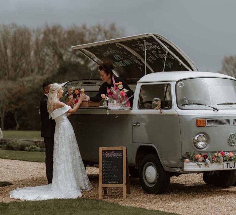 Groom in classic three piece grooms tuxedo with colourful spring boutonniere and bride in off the shoulder lace wedding dress getting ice cream from wedding food truck at Frensham Hall