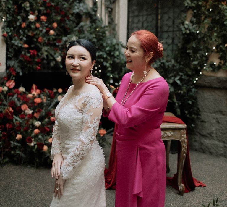 Mother of the bride wearing bright pink wedding outfit helping the bride into her wedding dress 