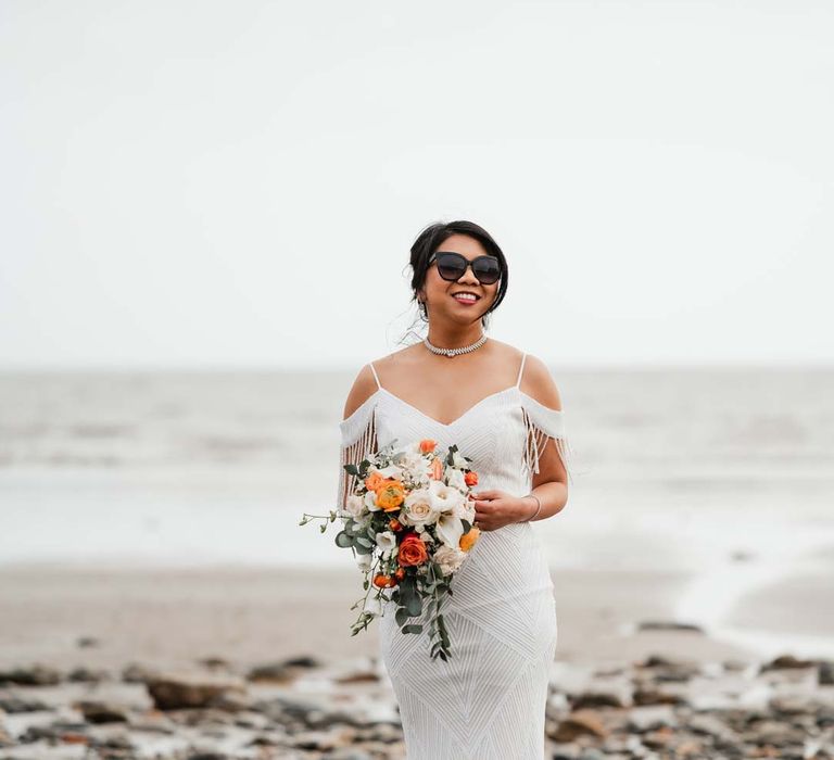 Bride in white beaded and sequined slip wedding dress holding mixed floral bouquet wearing wedding sunglasses at Welsh coastal wedding 