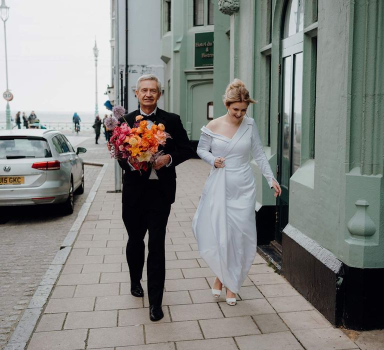 Bride in asymmetrical off shoulder long sleeve satin wedding dress walking with father of the bride in classic black suit in the streets of Brighton