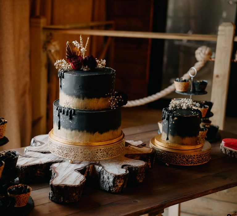 Gothic wedding dessert table with black wedding cupcakes, a two tier black wedding cake with gold rim and dried flower wedding cake toppers, and a single black wedding cake with gold rim and dried flower wedding cake toppers 