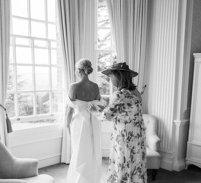 Mother of the bride in floral dress with hat helping the bride into her classic wedding dress 