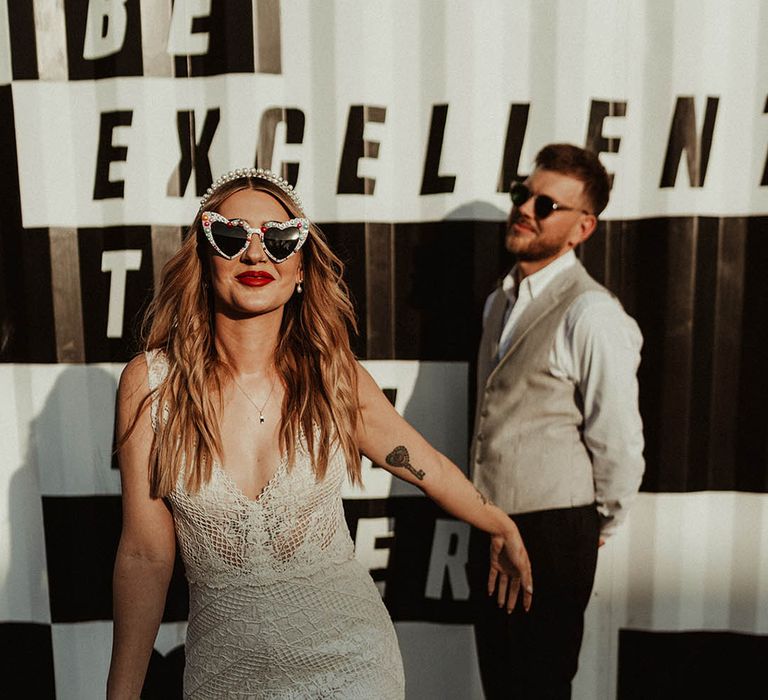 Bride wearing a lace wedding gown with red lipstick and retro sunglasses standing with the groom in a three piece suit 