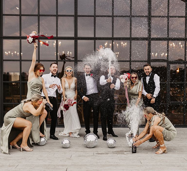 Champagne popping wedding photo outside the wedding venue with green bridesmaid dresses and disco balls