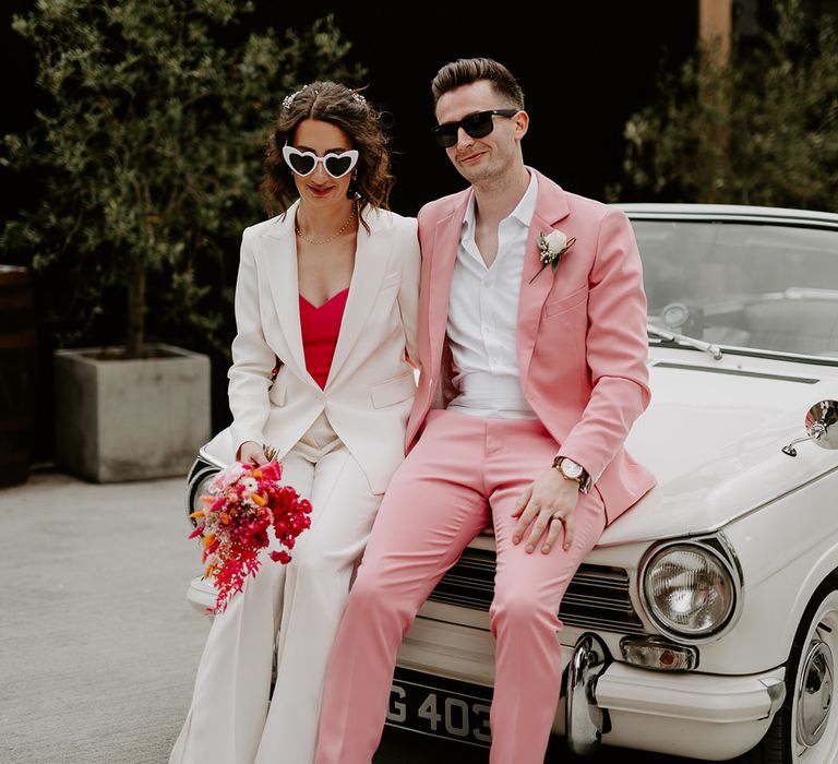 The Shack Revolution wedding with the bride and groom in pink and white wedding suits sitting on their vintage car 