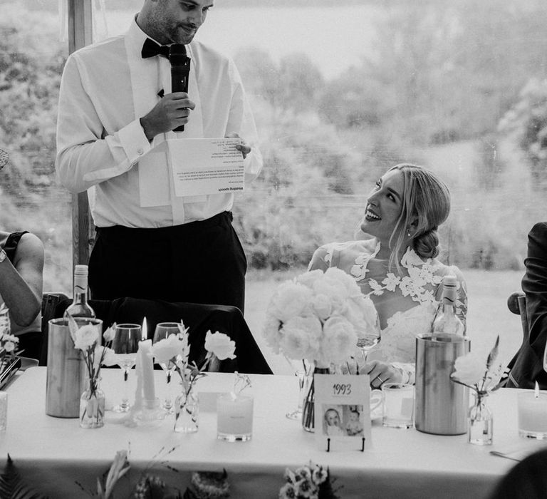 Groom reads out wedding speech at while bride looks up at him lovingly 