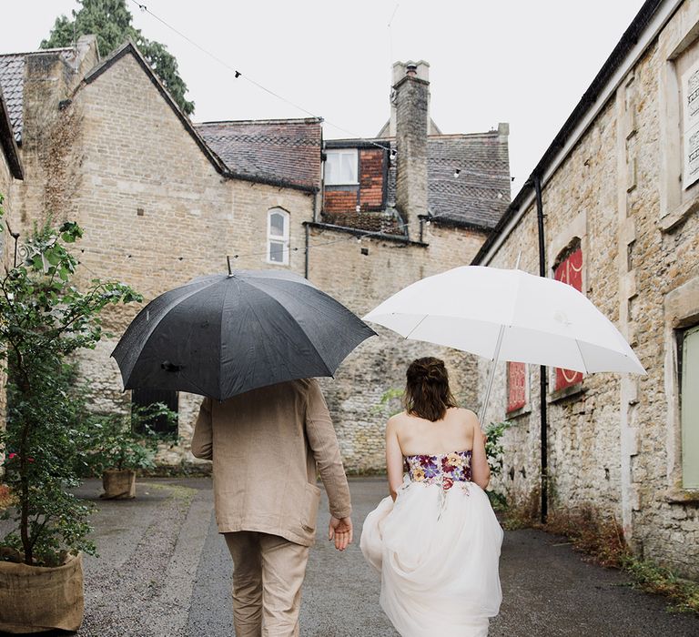 The bride and groom walking with umbrellas at rainy wedding in Somerset 