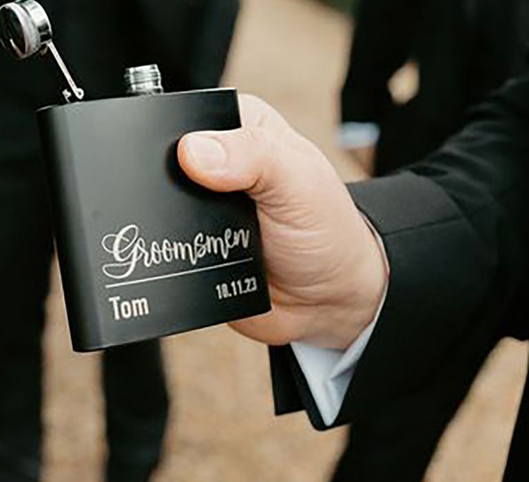 Groomsman holding out a black personalised flask with their name and the date of the wedding by Rachel Wood Photography