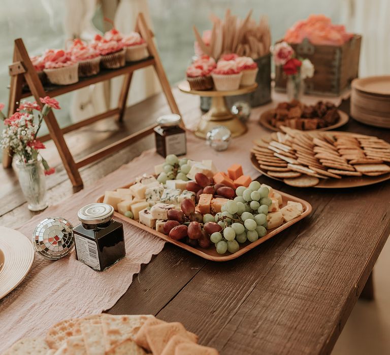 Charcuterie grazing table with crackers, fruit and jam with pink iced cupcakes 