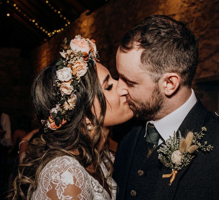 Boho bride with rose flower crown and curled wedding hair kisses groom in green tie and tartan at highland wedding in Glencoe