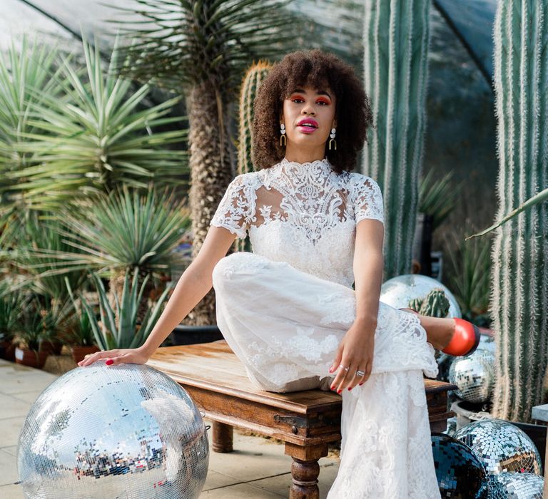 Bride in white jumpsuit and red heels sitting and resting hand on disco ball at Urban Jungle Suffolk