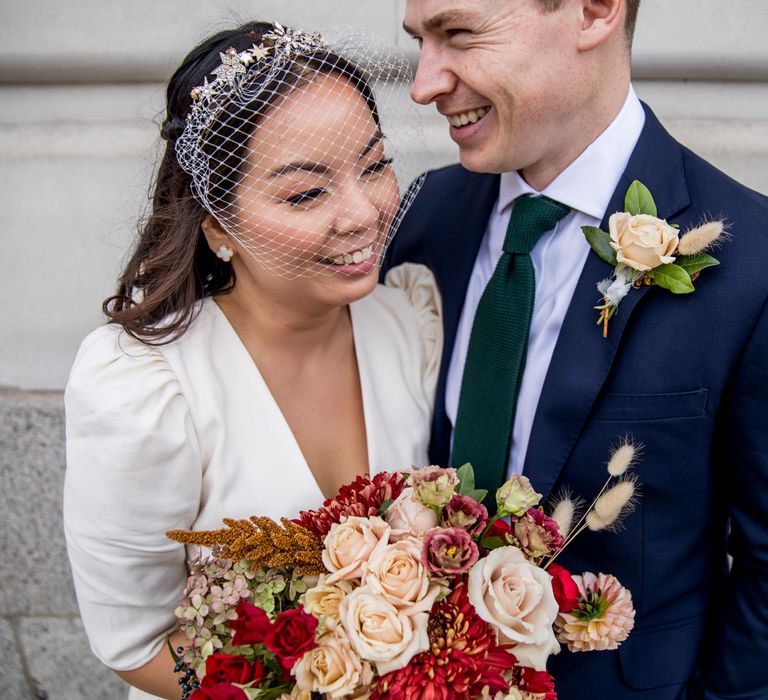 Asian bride in registry office wedding dress, birdcage veil and bridal crown laughing with her groom 