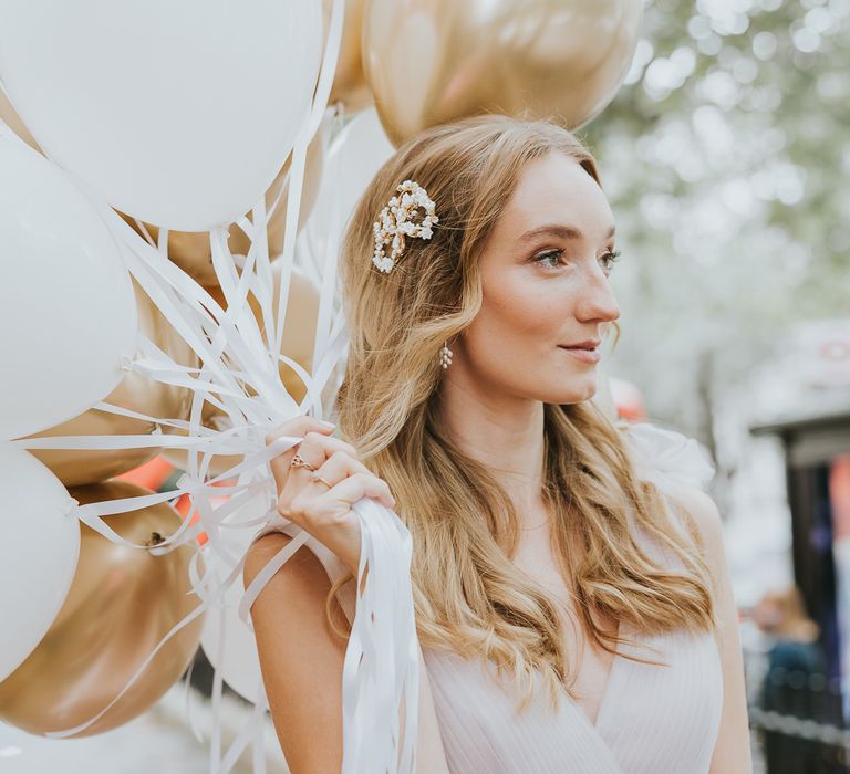 Bridesmaid with blonde wavy hair holding metallic biodegradable helium balloons wearing a gold and jewell encrusted hair slide 