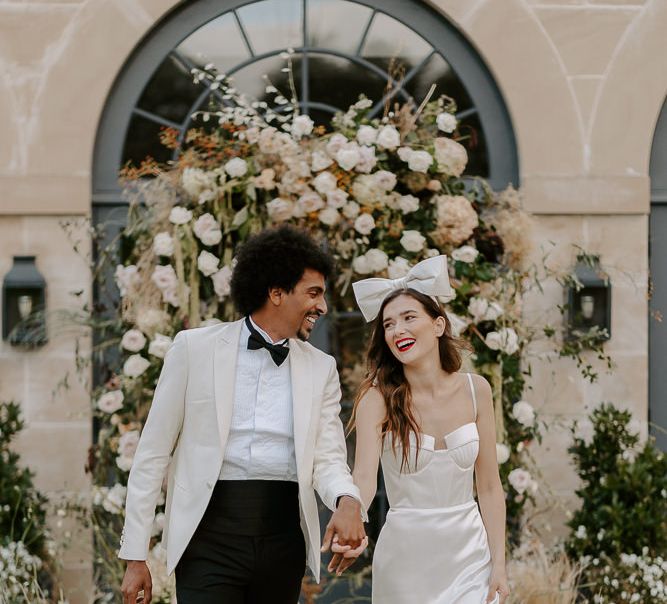 Bride in a satin wedding dress with cup detail and thin straps wearing a bow headdress laughing down the aisle with her groom in a white tuxedo jacket 