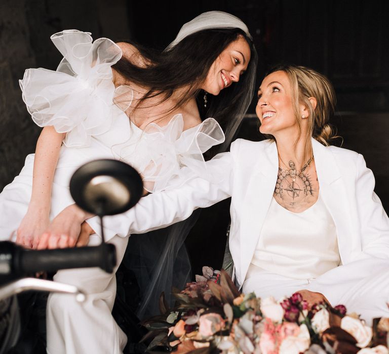 Bride in a satin top, trouser and jacket with chest tattoo kissing her bride 