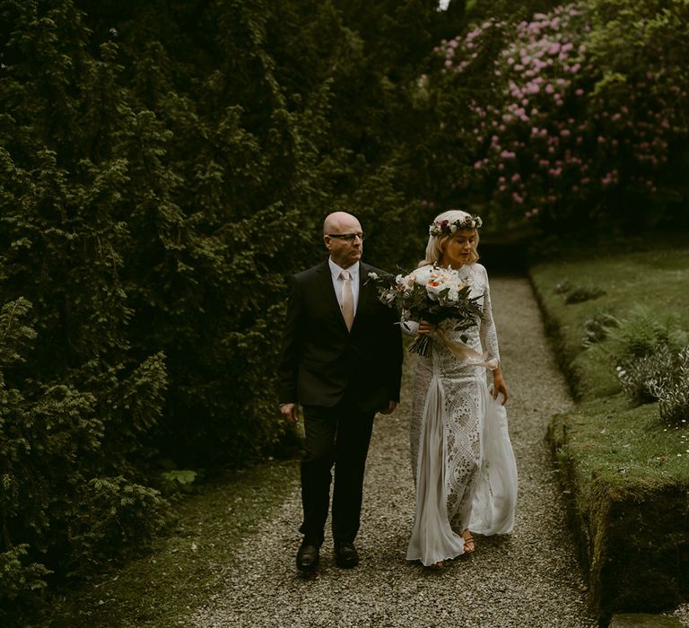 Bride in Grace Loves Lace Wedding Dress, single tier cathedral length veil and flower crown holding pink and white rose bouquet walking with father in the gardens