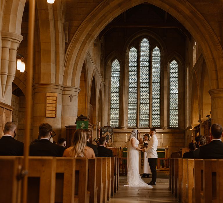 Groom in white tuxedo jacket holds hands with bride in white Made With Love wedding dress and white chapel length veil at the church altar