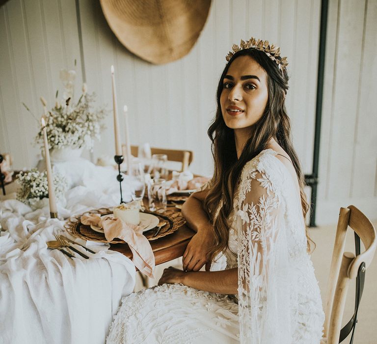 Boho bride in a lace wedding dress and gold crown sitting at the table decorated with dried flowers and taper candles in neutral tones 