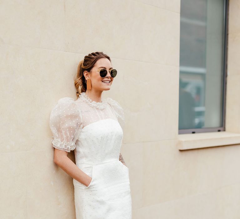 Bride in a knee length polka dot wedding dress with puff sleeves and sunglasses leaning up against the wall 