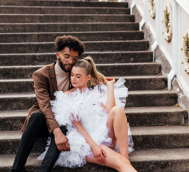 Groom in a brown polo neck and blazer embracing his bride on the steps in a layered tulle wedding dress 