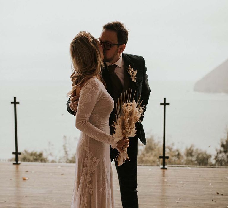 Bride in a fitted wedding dress with floral design holding a dried flower bouquet kissing her groom during the wedding ceremony 