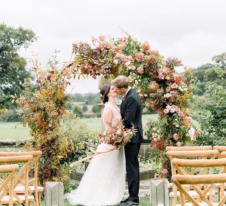 Groom in a tuxedo kissing his bride in a tulle wedding dress in front of a large floral arch