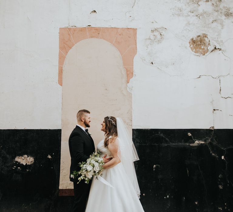 Bride & groom face one another and look lovingly whilst smiling in front of wall outdoors in London