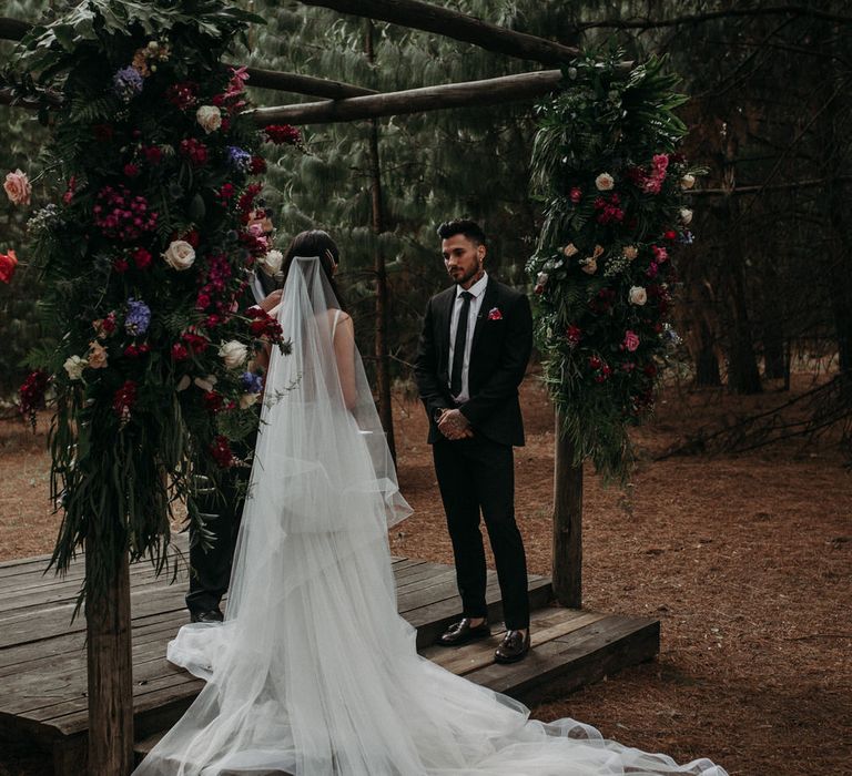 Bride and groom getting married under moody romantic wedding flowers in outdoor woodland ceremony