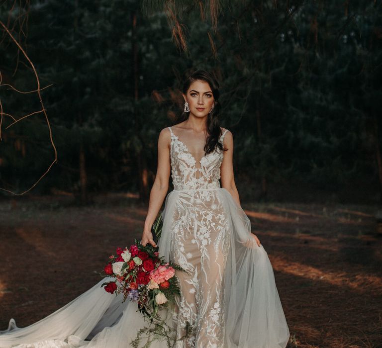 Bride wearing a lace embroidered wedding dress with tulle overskirt, carrying a bouquet of red and pink peonies and roses