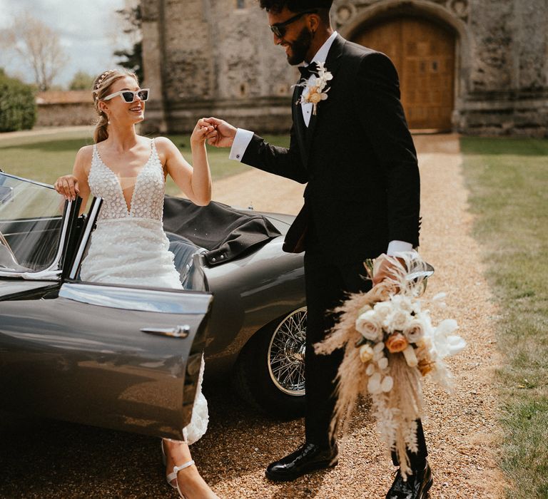 Groom in a tuxedo helping his bride in an appliqué wedding dress with deep V front out of their vintage wedding car 