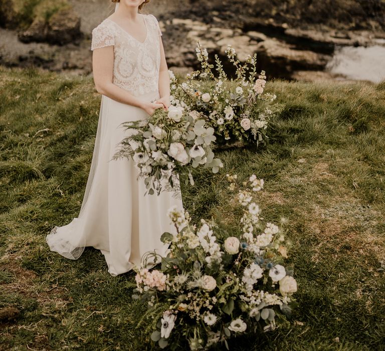 Bride in lace top wedding dress with capped sleeves and satin skirt holds mixed wedding bouquet with white and green florals whilst standing on clifftop with Dunluce Castle in the background