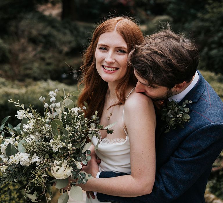 Groom in a navy blue suit kissing his brides shoulder in a slip wedding dress as she holds her green foliage and white flower bouquet 