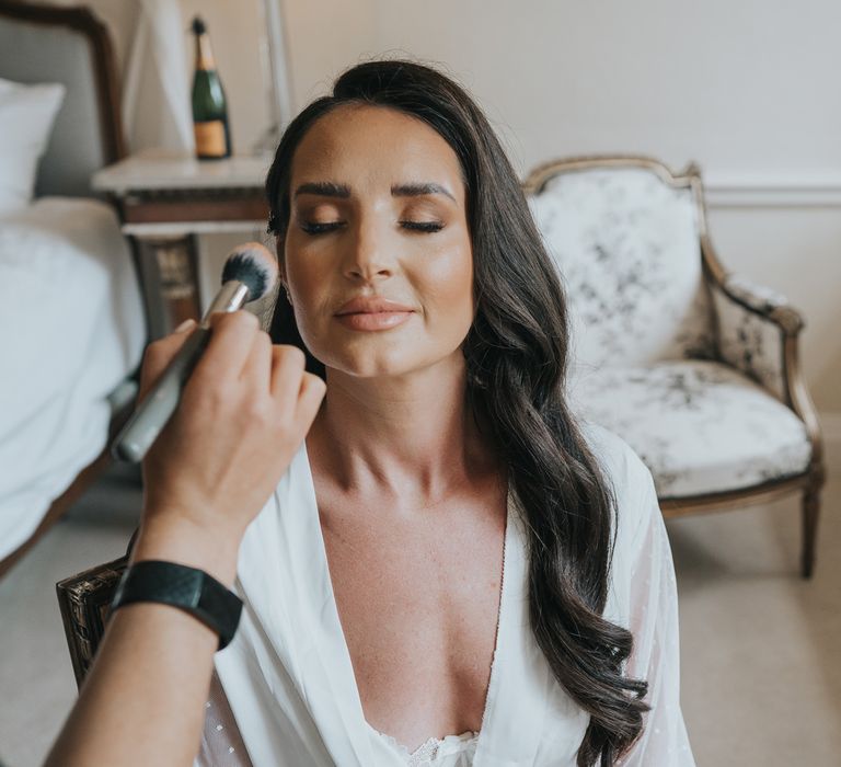 Bride with long dark curled hair in white satin bridal dressing gown has her make up done in bedroom at Came House Dorset