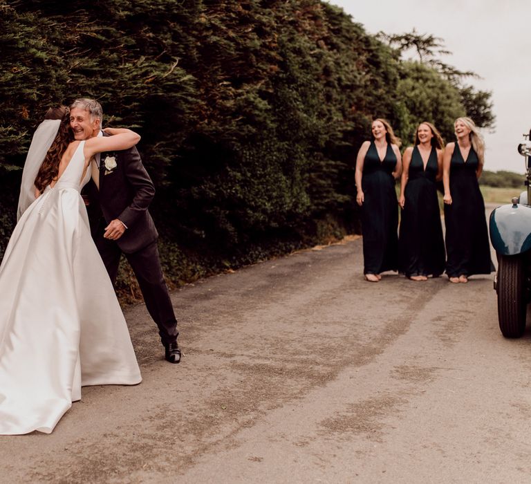 Bride in low back Elbeth Gillis wedding gown with train and veil hugs father whilst bridesmaids in green dresses watch