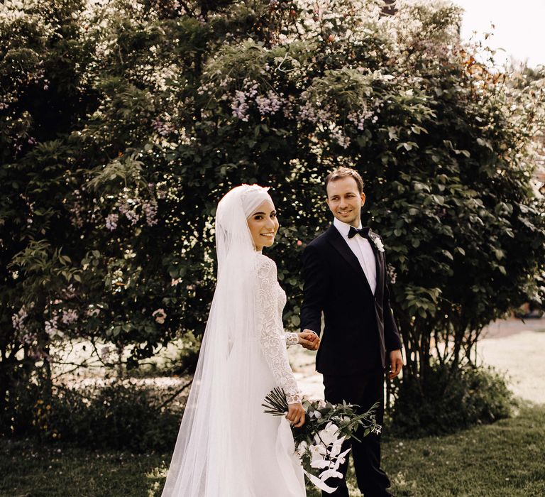 Bride in high neck long sleeve 3D lace wedding dress with puddle train holding hands with groom in black tie