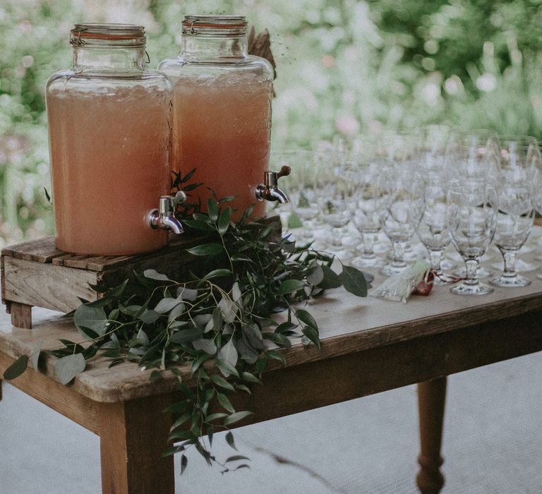 Two large glass drinks dispensers with ice and orange drink stand on wooden table with green foliage decor and glassware under tipi at garden wedding with burnt orange wedding theme