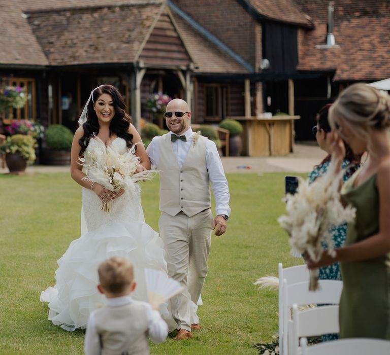 Bride in a Mori Lee mermaid wedding dress walking down the aisle being greeted by her son in a beige suit 