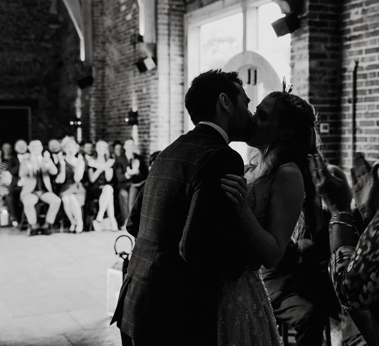 Bride in pearl wedding dress and groom in checked suit kiss during barn wedding ceremony