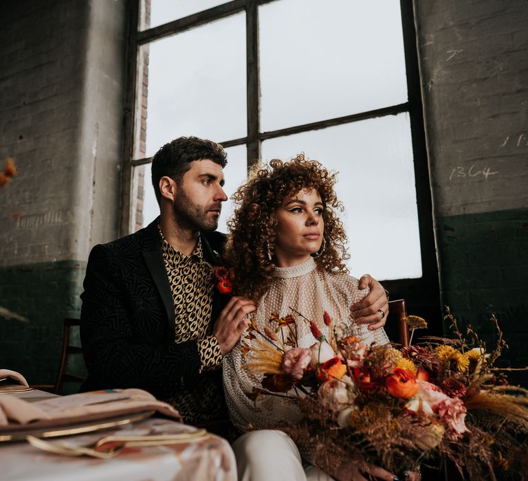 Bride in a jumpsuit and groom in a black and gold patterned shirt sitting at their intimate tables ape decorated with dried and fresh flowers and coloured glassware 