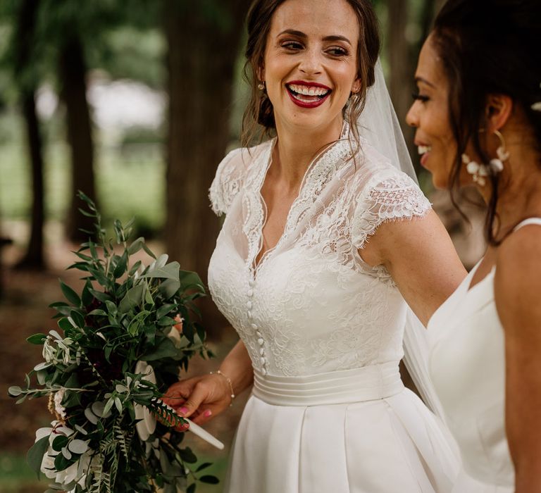 Laughing bride in white lace top capped sleeve wedding dress with bold red lip holding bridal bouquet looks at bride with curled ponytail in white cami wedding dress with shoulder veils after wedding ceremony at The West Mill Derby