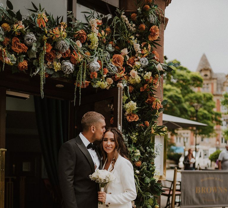 Groom in a black tuxedo kissing his bride's forehead in a doorway decorated with flowers 