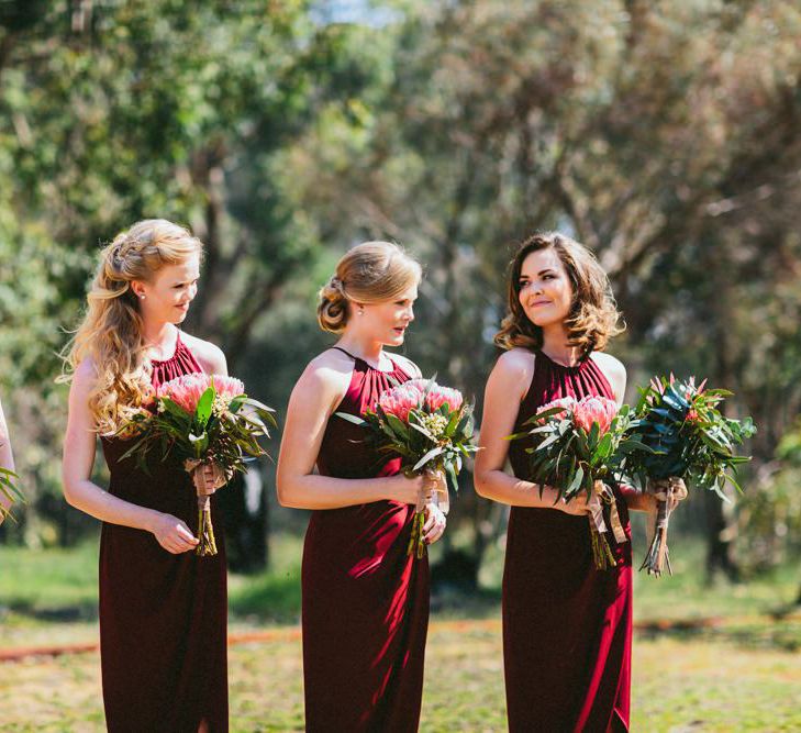 Bridesmaids standing at the outdoor wedding ceremony in halter neck burgundy dresses holding pink protea bouquets 