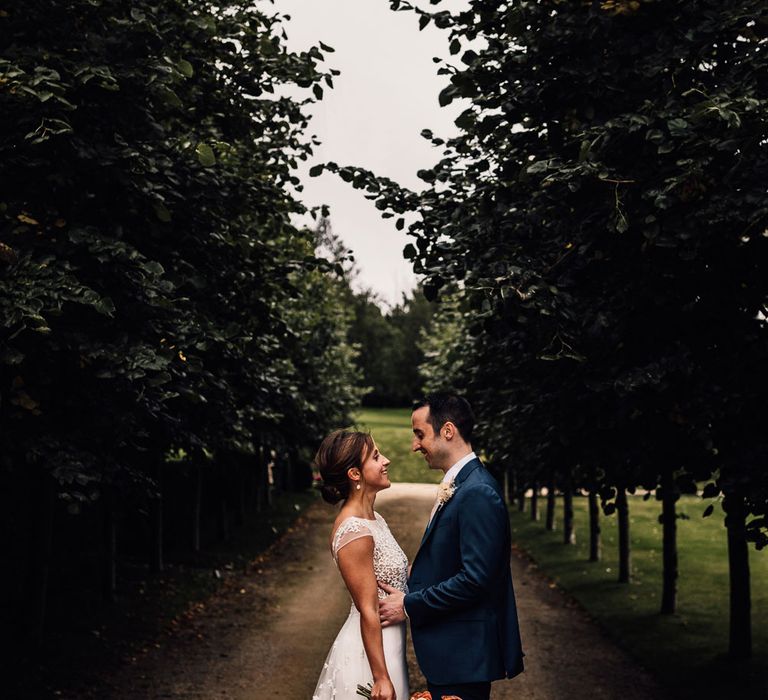 Bride in white open back Rime Arodaky wedding dress with lace train holding dried bridal bouquet smiles at groom in blue suit as they stand in driveway after summer wedding ceremony in Dorset