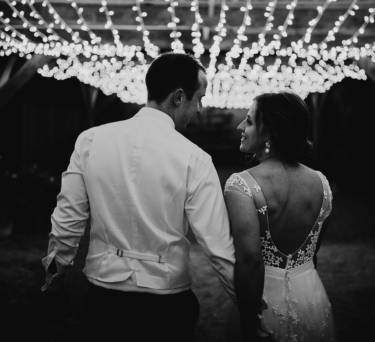 Bride in open back Rime Arodaky wedding dress walks holding hands with groom in waistcoat under fairy light canopy at summer wedding in Dorset